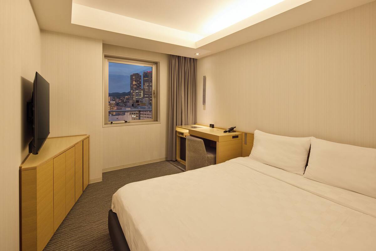 Een hotelkamer met een bed, een bureau, een televisie en een raam met uitzicht op de stad.