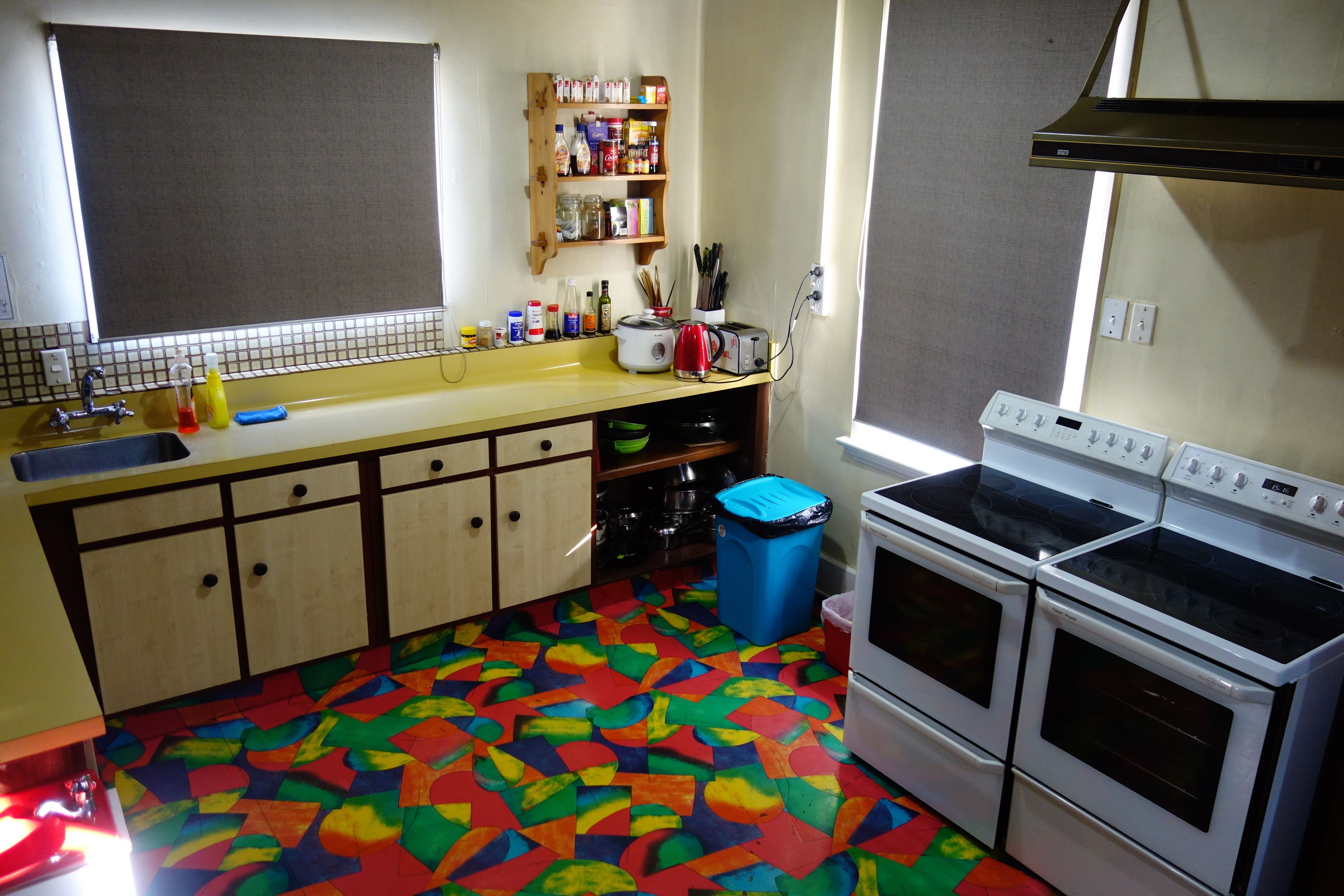 Een keuken met een vloer in verschillende kleuren, een spoelbak, kastjes, een fornuis en een raam.
