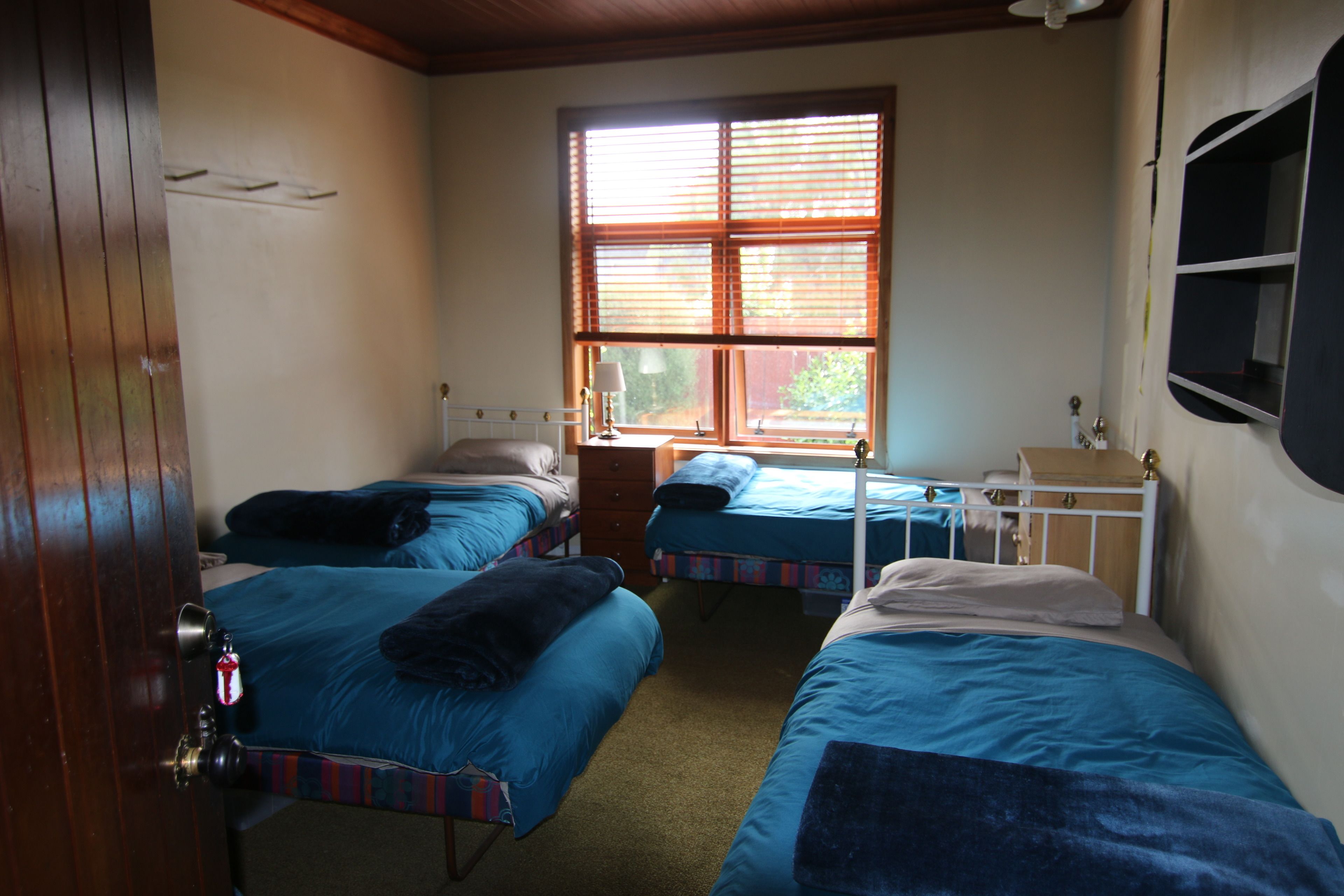 A room with three single beds, a wooden door, a window with blinds, and a small bedside table with a lamp.