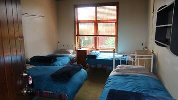 A room with three single beds, a wooden door, a window with blinds, and a small bedside table with a lamp.