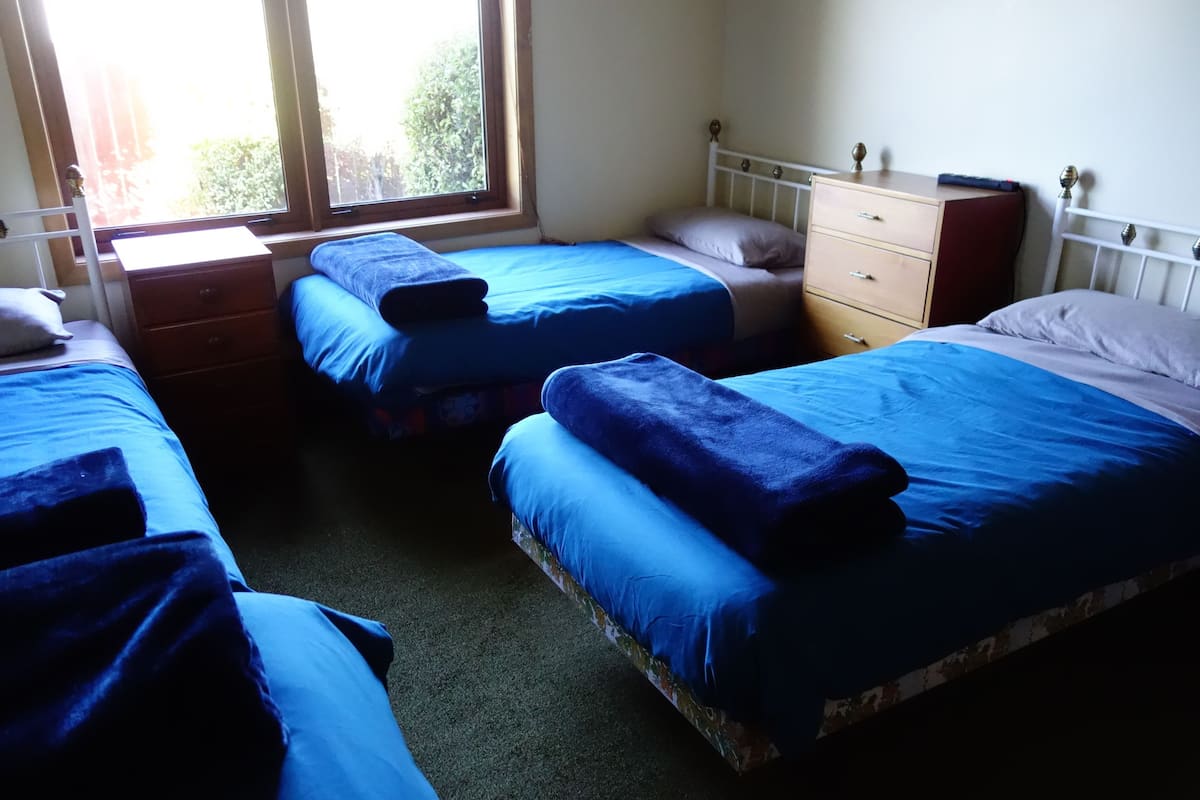 Una habitación con tres camas individuales, cada una con una manta y almohada azules. Hay un aparador de madera con cajones y un espejo. Un ventanal proporciona luz natural.