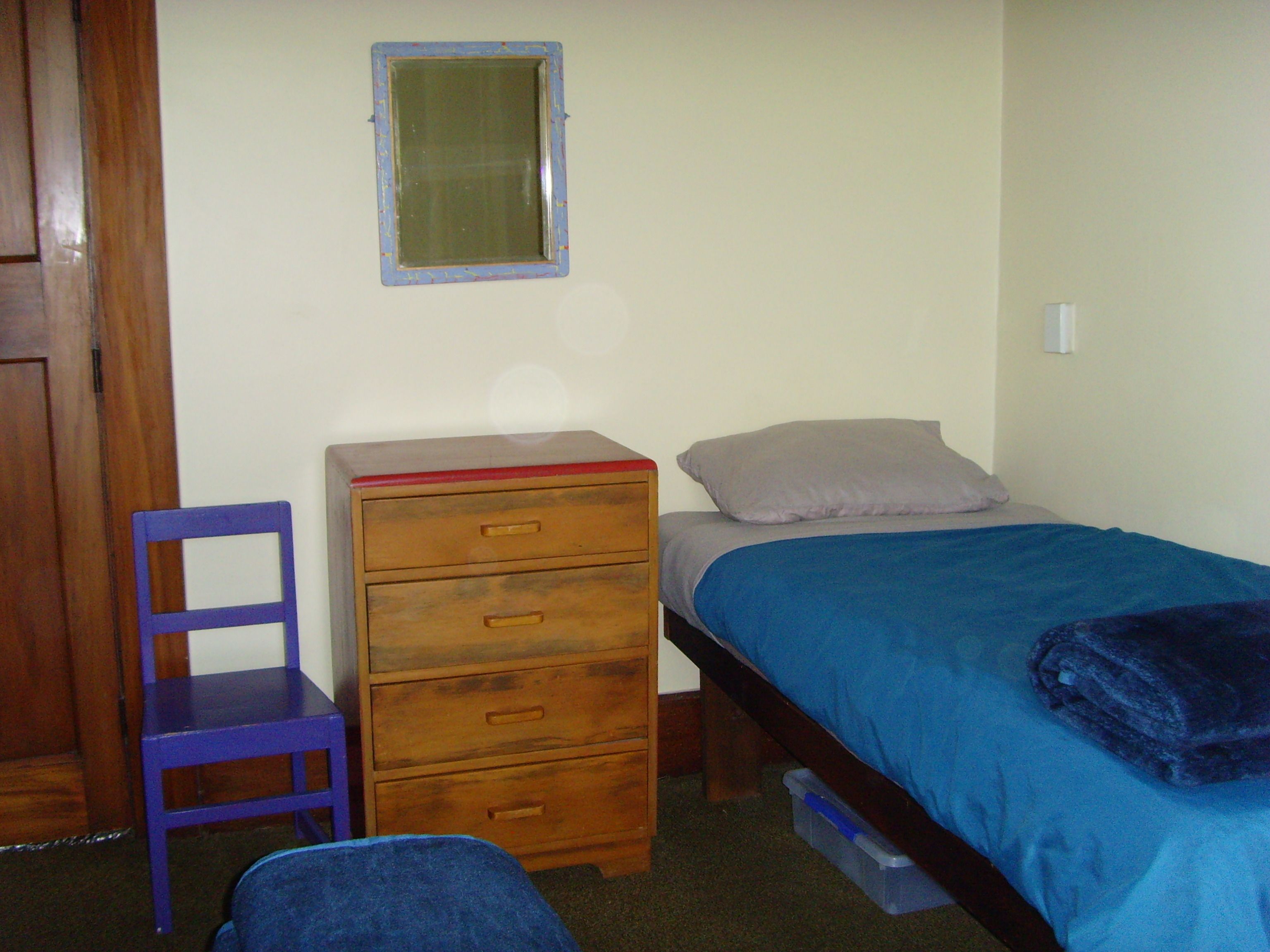 Une petite chambre avec un lit simple, une commode en bois, une chaise et une fenêtre.