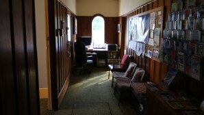 A hallway with a wooden floor, a desk with a computer, a chair, and a bulletin board with papers and notices. - Big Tree Lodge Methven - Hostel (Methven)