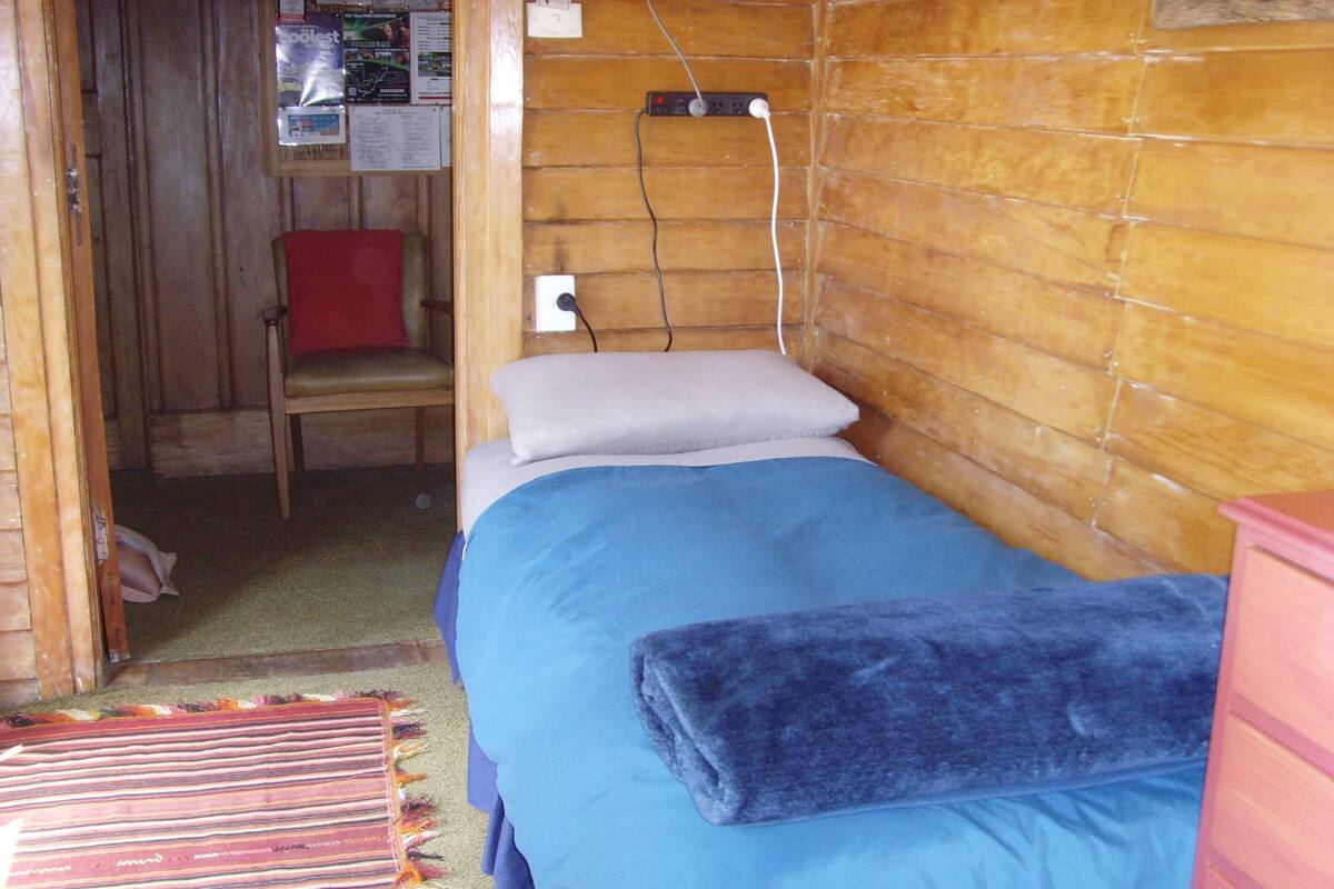 Una habitación pequeña con una cama individual, cabecera de madera, una silla roja y un enchufe en la pared.