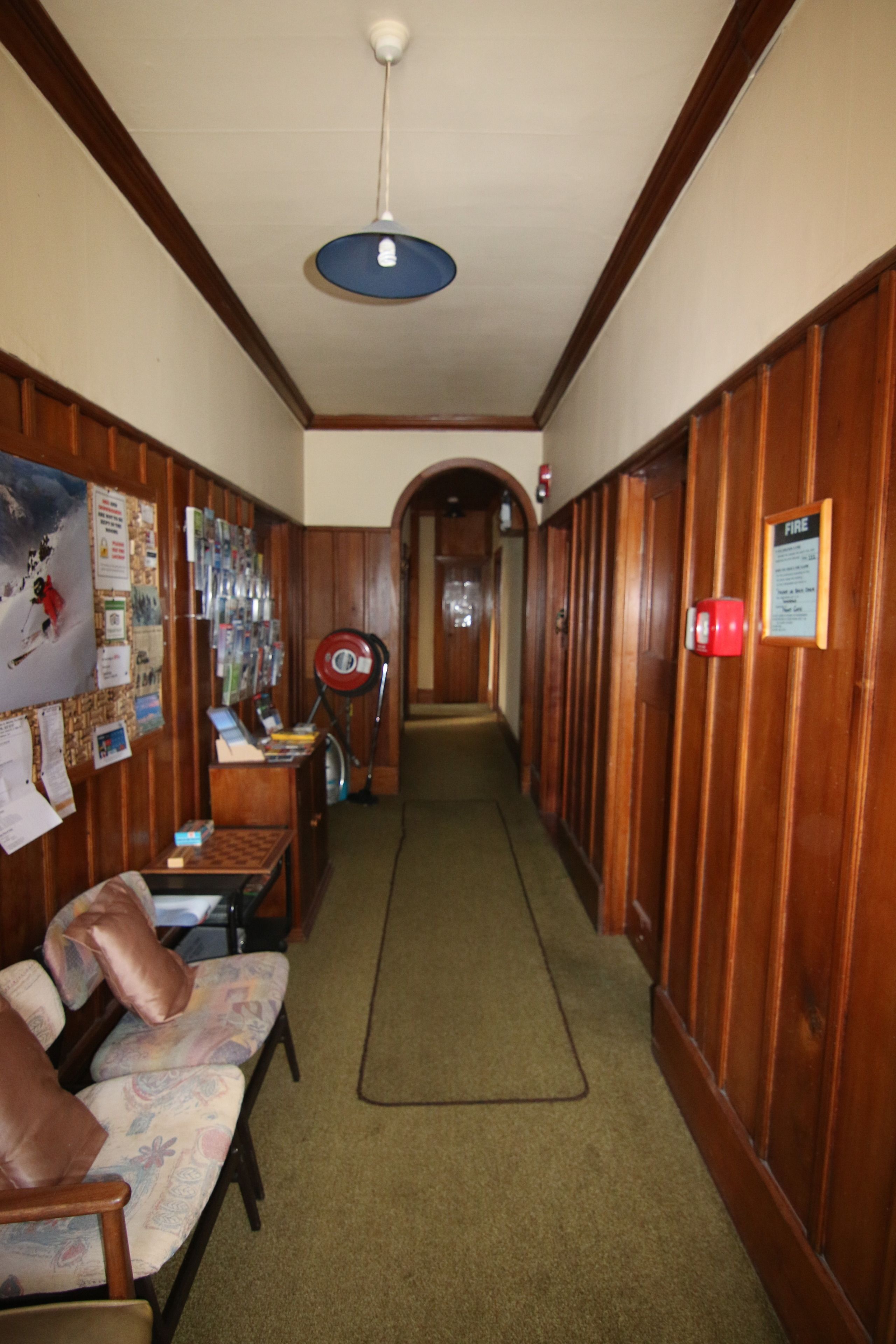 A hallway with wooden paneling, a green carpet, a blue ceiling light, a fire alarm, and a bulletin board with various notices.