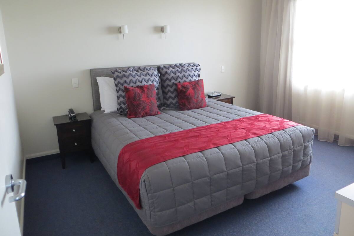 A neatly made bed with a grey quilt and red accents, a bedside table with a phone, and a window with sheer curtains.