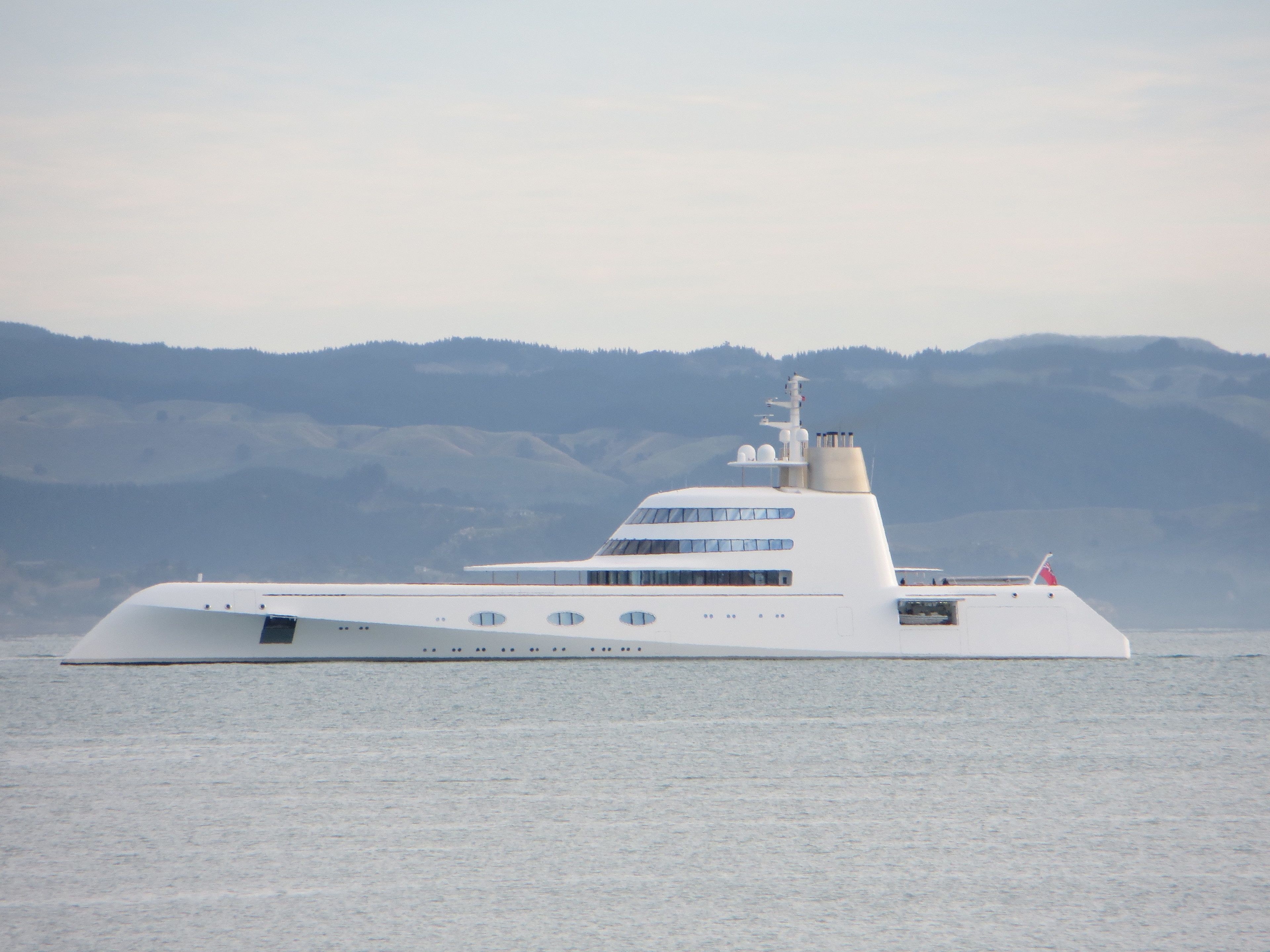 A large, modern yacht with multiple decks and a sleek design, floating on calm waters with a mountainous backdrop.