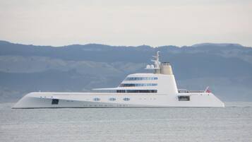 A large, modern yacht with multiple decks and a sleek design, floating on calm waters with a mountainous backdrop.