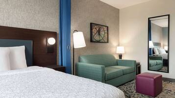 A hotel room with a large bed, a green armchair, a small table, a mirror, and a wall clock.