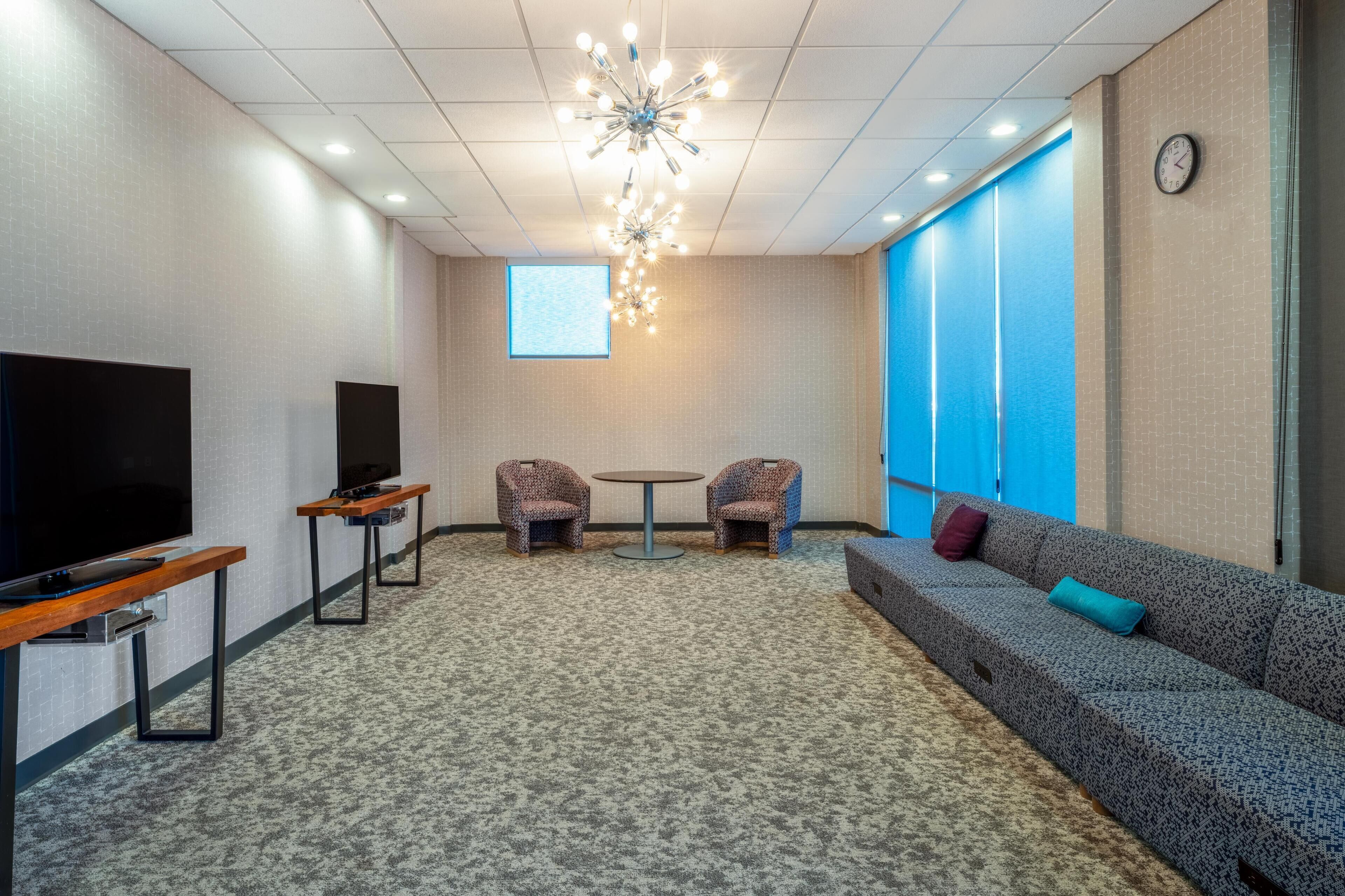 A conference room with a large flat-screen TV, two patterned chairs, a round table, a sofa with cushions, a ceiling light fixture, and a clock on the wall.
