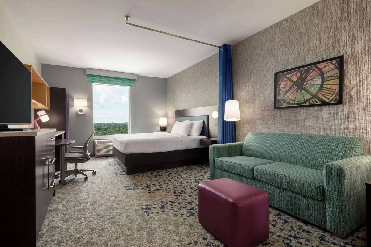A hotel room with a bed, a green sofa, a desk with a chair, and a large window offering a view.
