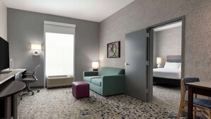 A hotel room with a desk, a green armchair, a small purple ottoman, a bed with a white headboard, and a large window. - Home2 Suites by Hilton Florence, SC (Florence)