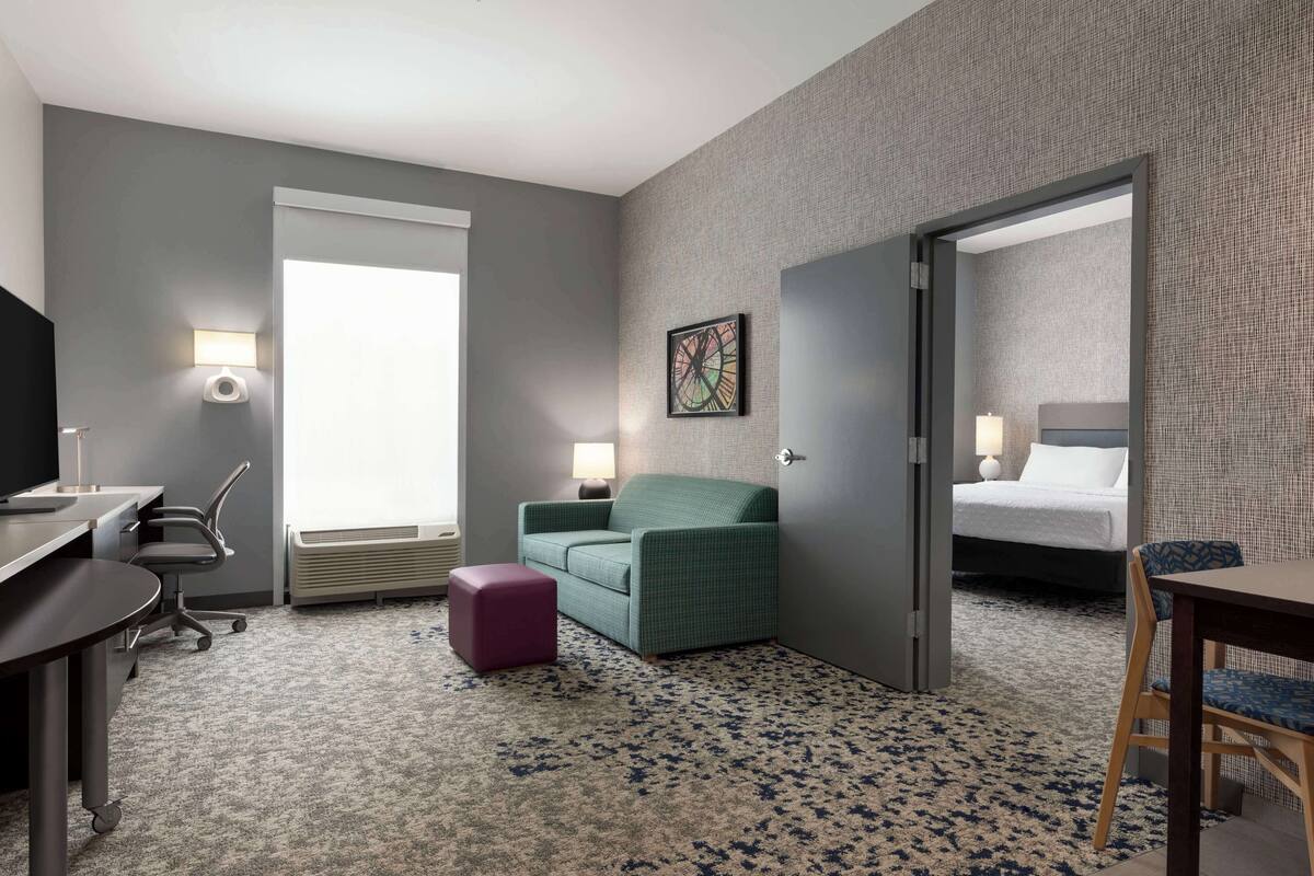 A hotel room with a desk, a green armchair, a small purple ottoman, a bed with a white headboard, and a large window.
