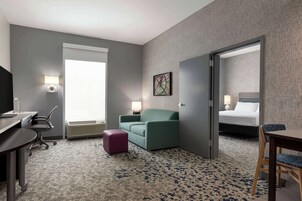 Habitación de hotel con escritorio, un sillón verde, un pequeño taburete morado, cama con cabecera blanca y un ventanal grande.