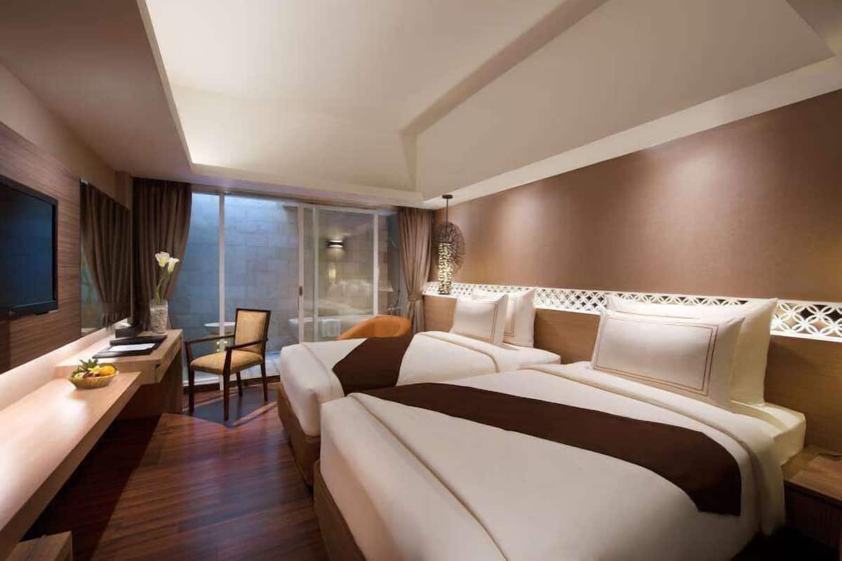 Habitación de hotel con una cama grande, un escritorio y una silla. Se aprecia una ventana grande con cortinas, y un adorno decorativo sobre la cama.