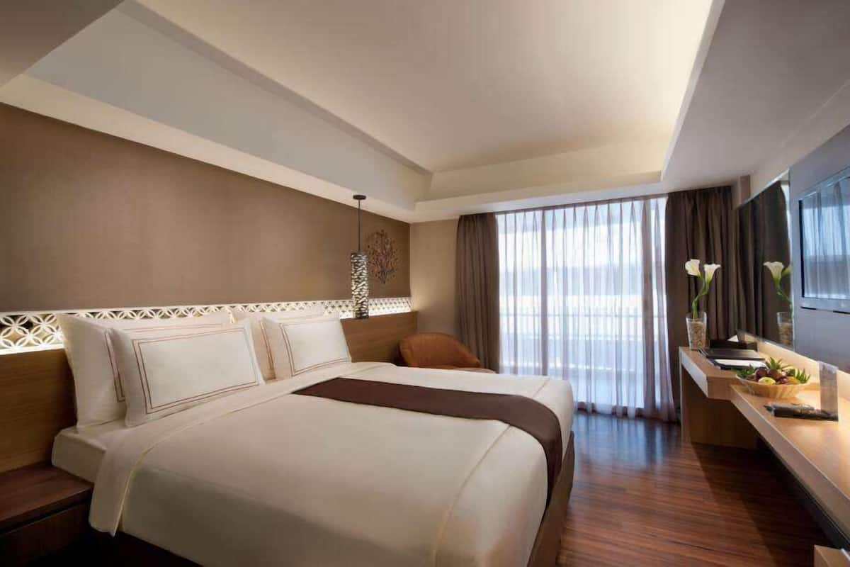 Habitación de hotel con una cama grande, un escritorio con un florero y frutas, y vistas al exterior a través de cortinas translúcidas.