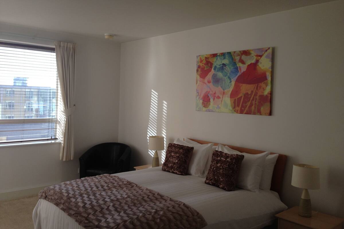 Habitación de hotel con cama, una silla, mesita de noche, lámpara y ventana con cortinas.