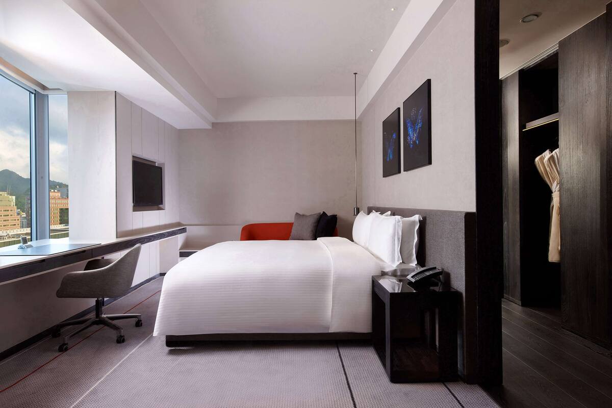 A modern hotel room with a large bed, a desk, a chair, and a view of the city.