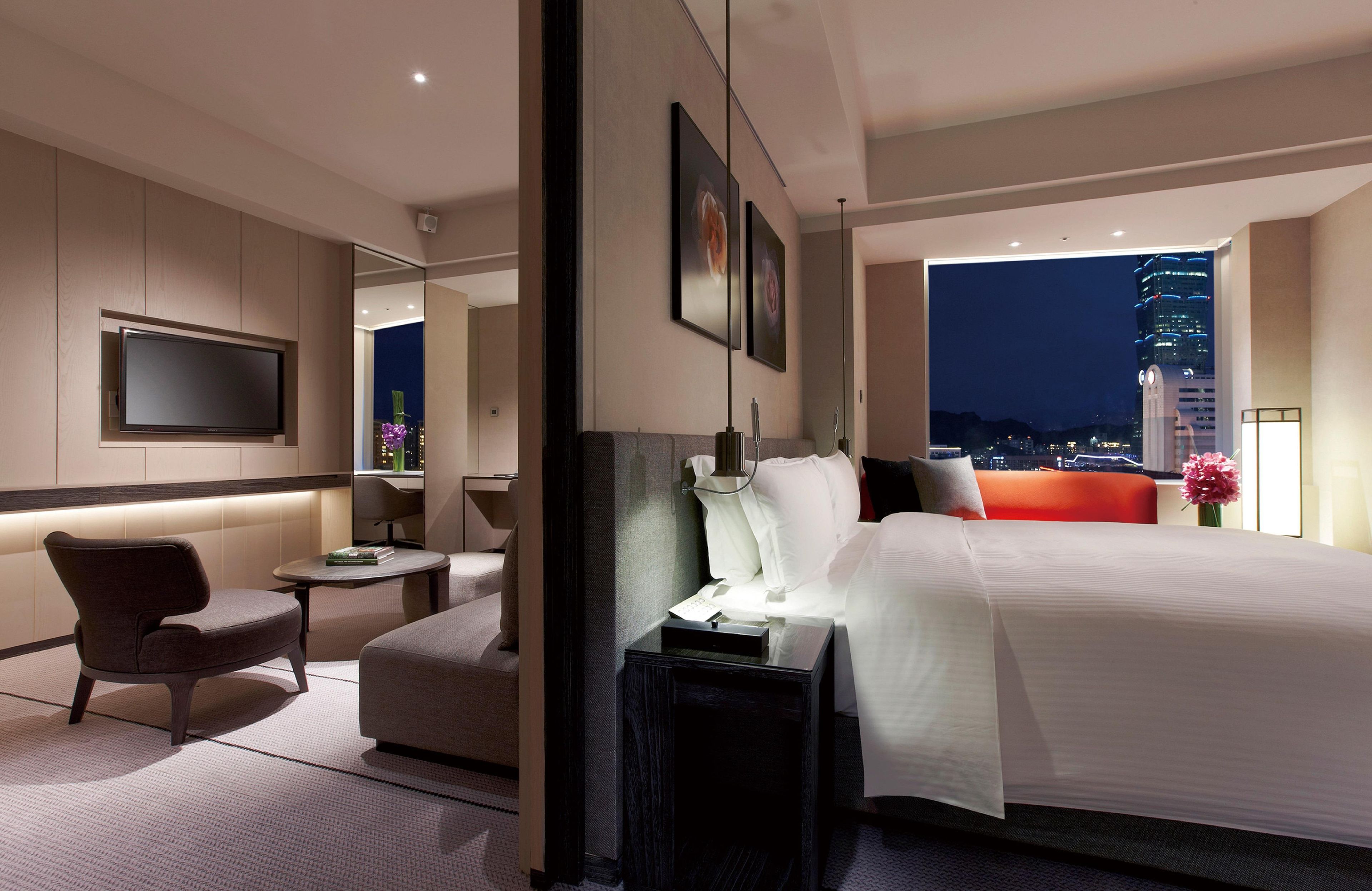 A modern hotel room with a large bed, a seating area, a flat-screen TV, and a city view.