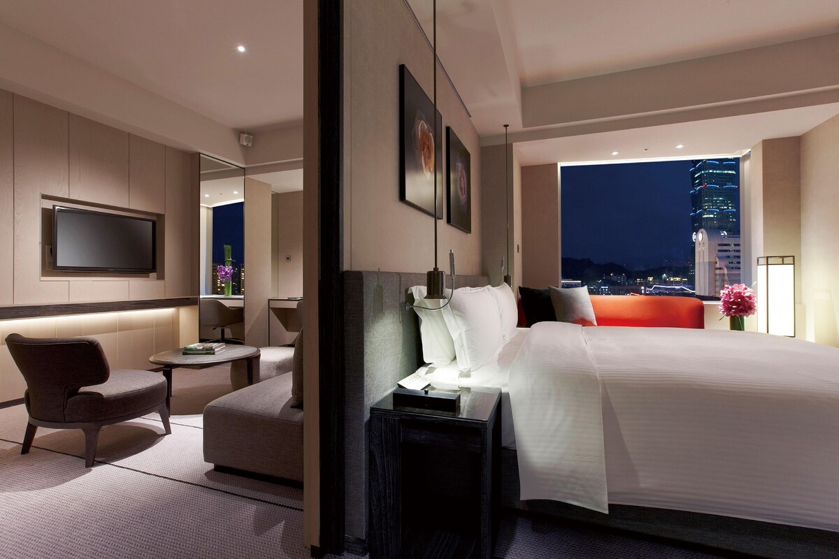 A modern hotel room with a large bed, a seating area, a flat-screen TV, and a city view.