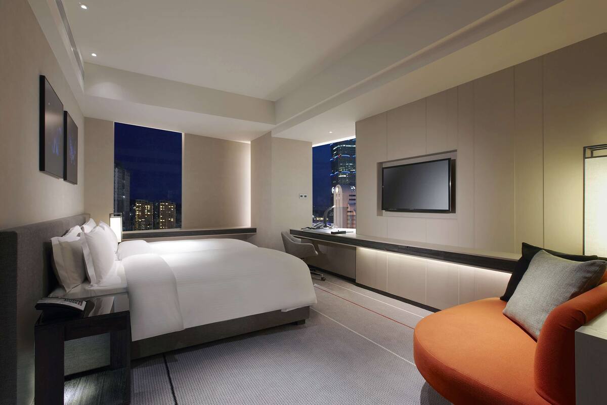 A modern hotel room with a bed, a desk, a chair, and a TV on the wall.