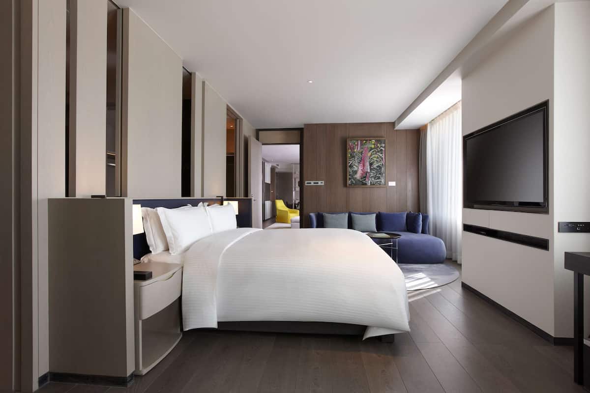 A modern hotel room with a large bed, a TV, a desk, and a seating area.