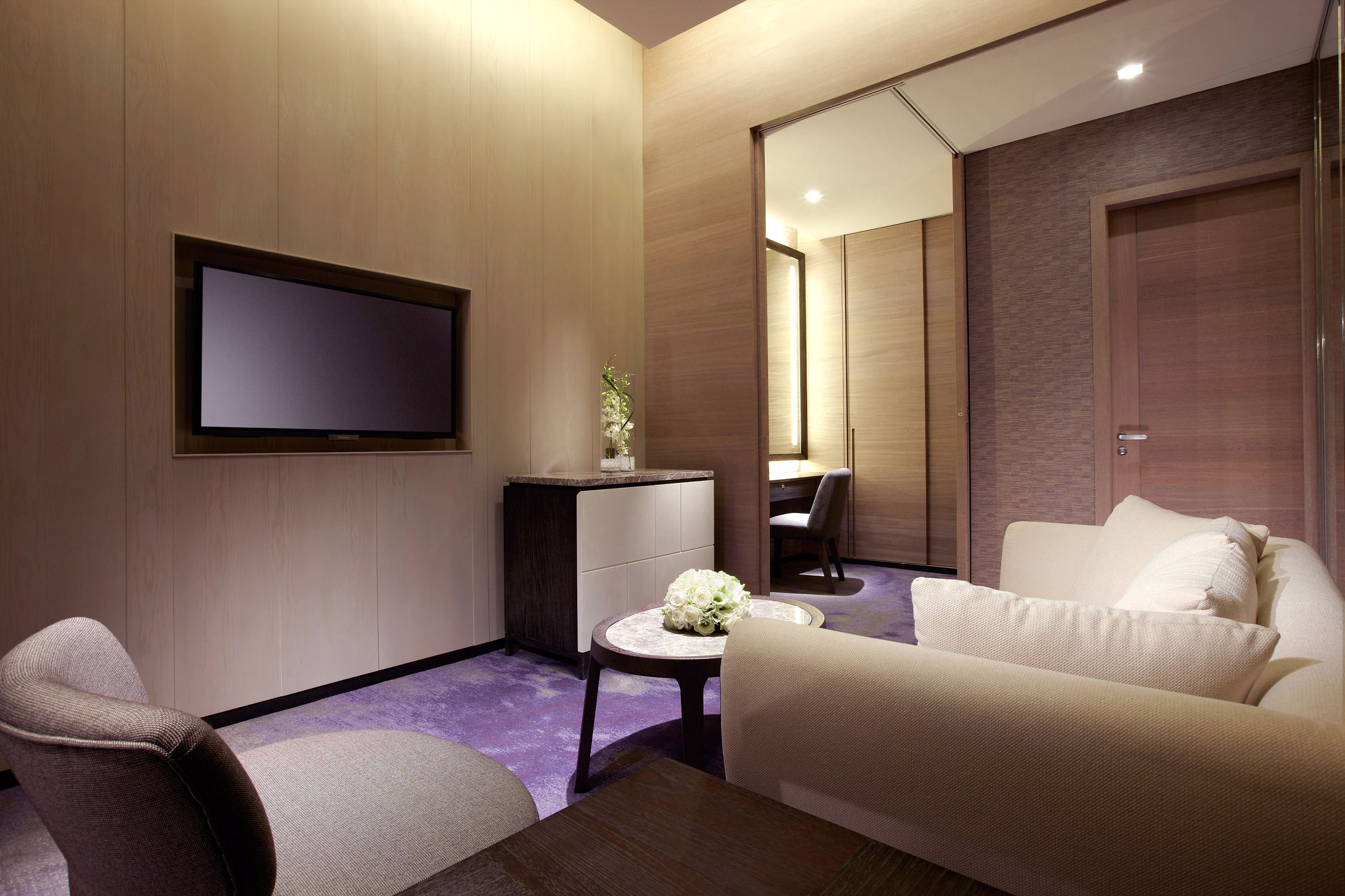 A modern hotel room with a flat-screen TV, a small round table with a vase of flowers, a sofa, and a chair.