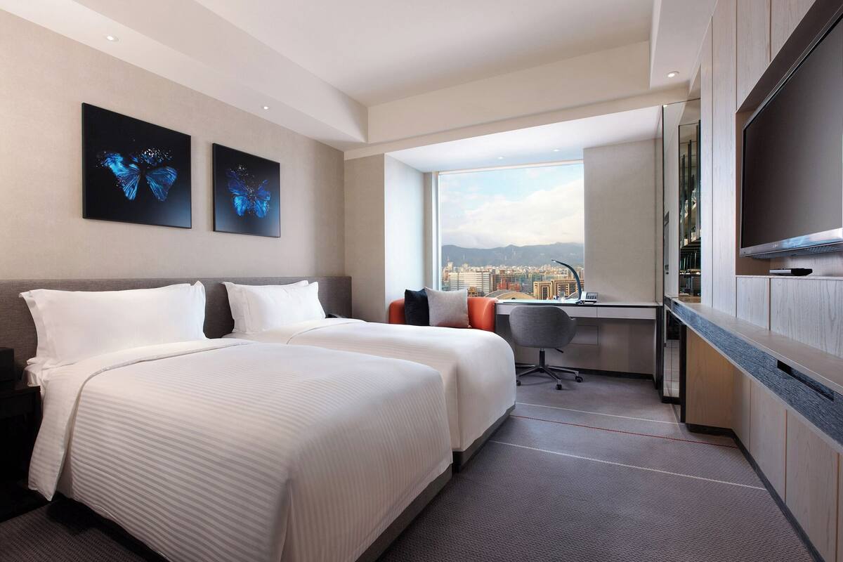 A hotel room with two beds, a desk, a TV, and a city view.