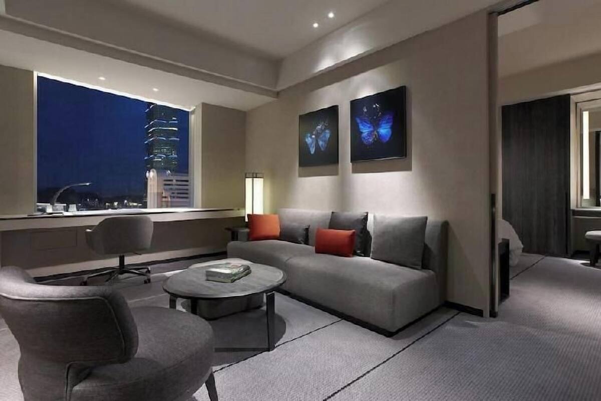 A modern hotel room with a sofa, armchairs, a coffee table, and a large window with a city view.