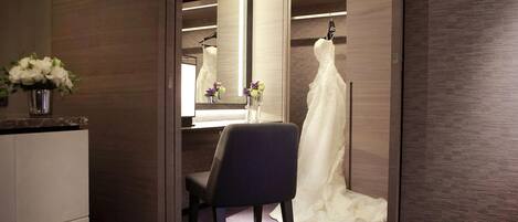 A hotel room with a walk-in closet, a vanity with a mirror, and a wedding dress on a hanger.