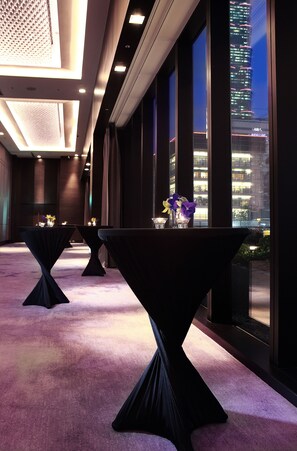 Un hall de hotel con mesas y sillas, amplios ventanales con vistas a la ciudad y suelo alfombrado.