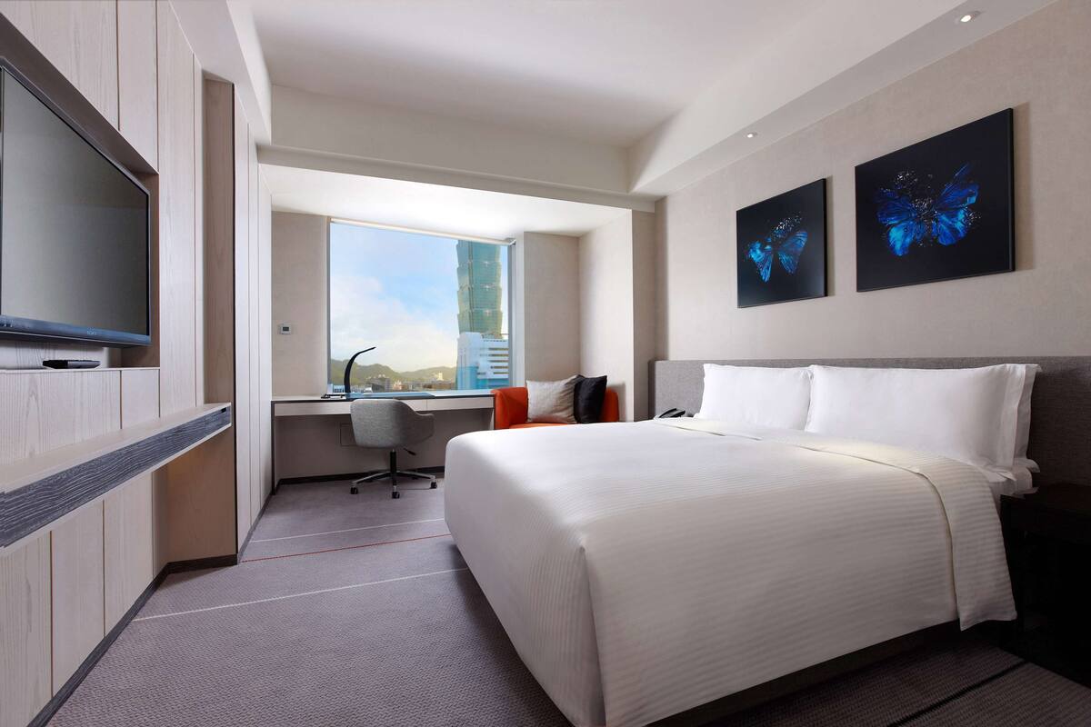 A hotel room with a bed, a desk, a chair, a TV, and two butterfly paintings on the wall.
