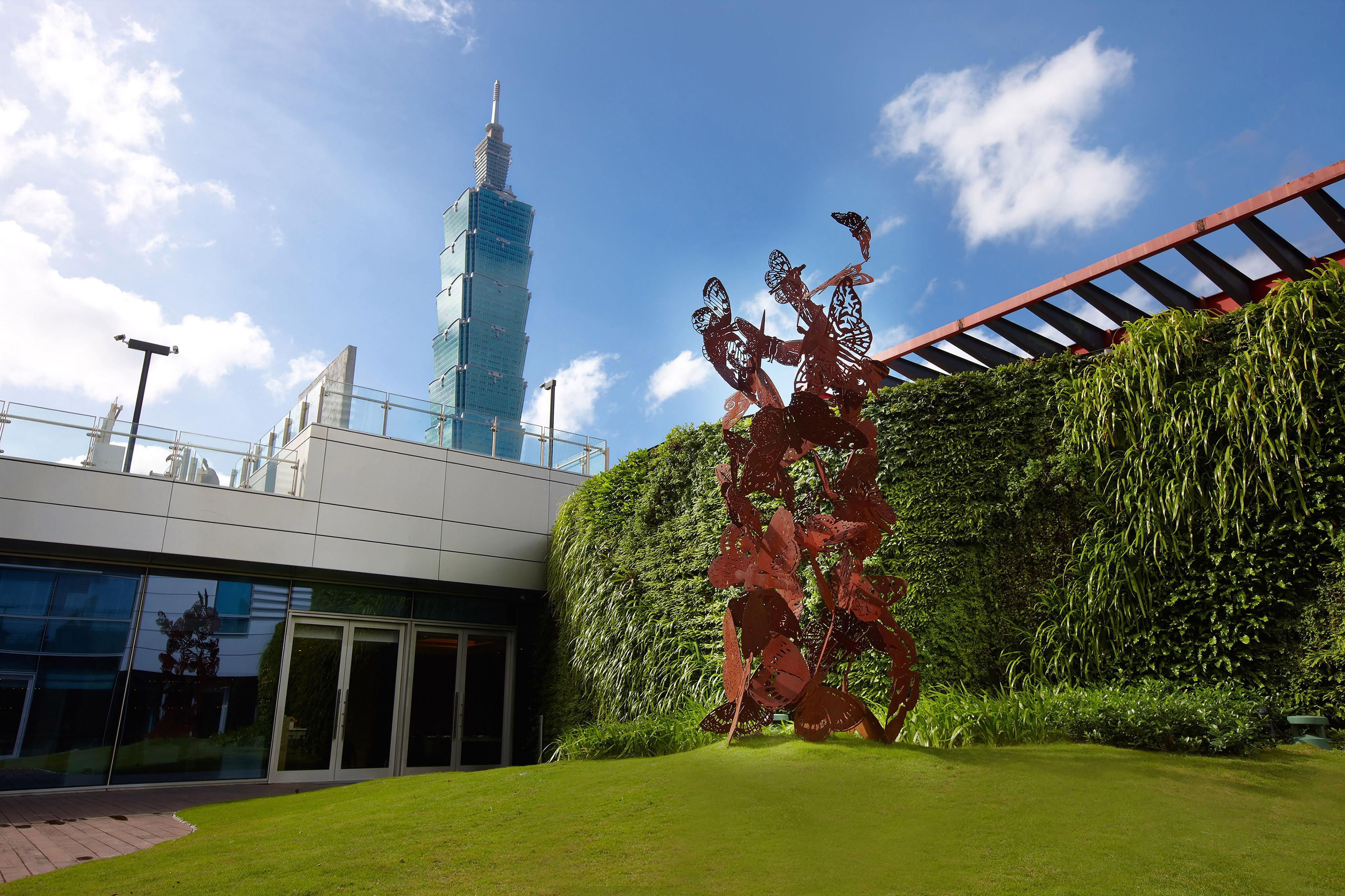 A modern building with a glass facade, a metal sculpture in the foreground, and a tall tower in the background.
