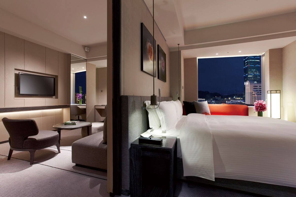 A modern hotel room with a large bed, a TV mounted on the wall, a seating area with a chair and a small table, and a view of a cityscape at night.