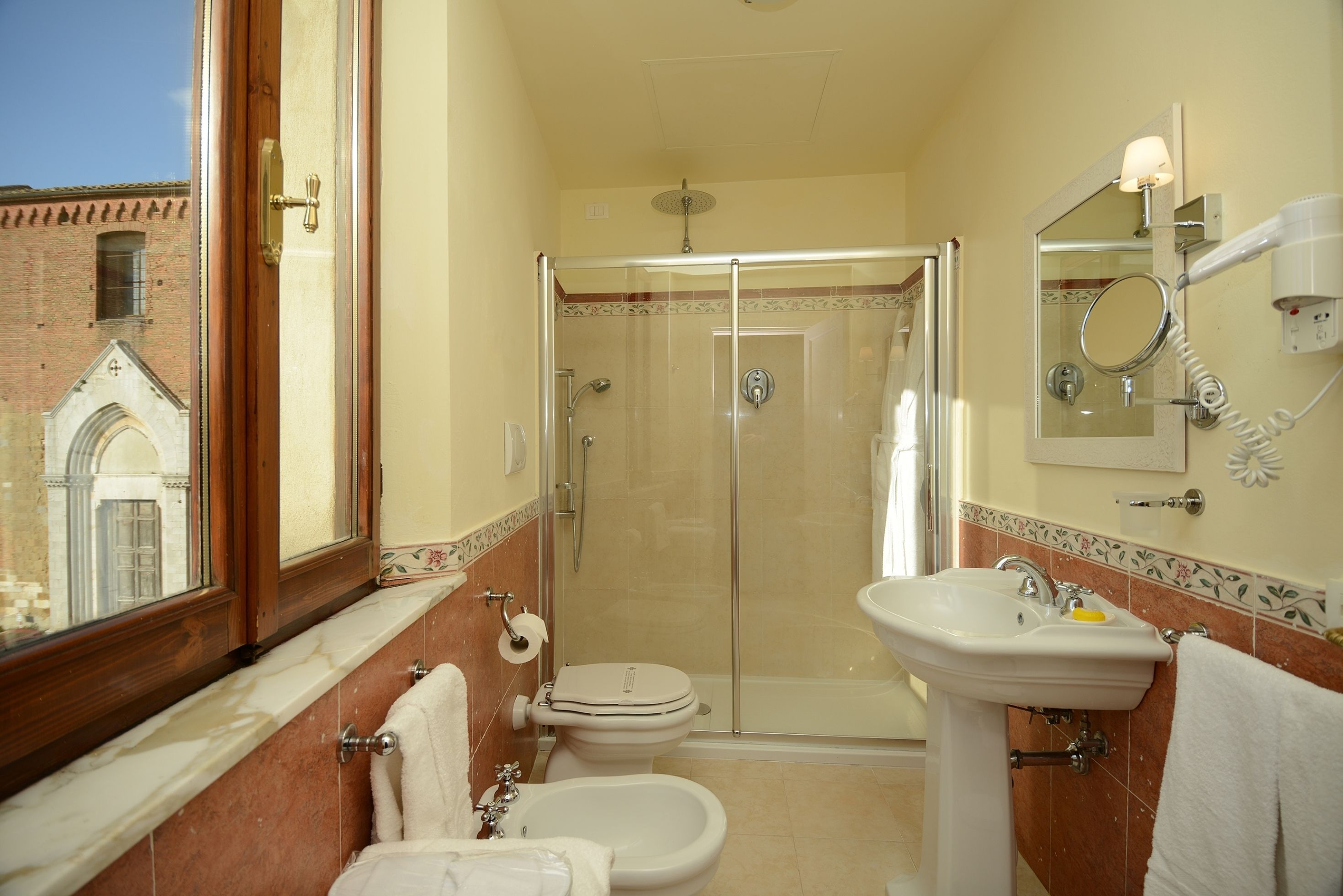 Een badkamer met douche, toilet, bidet, wastafel en een raam met uitzicht op een gebouw.