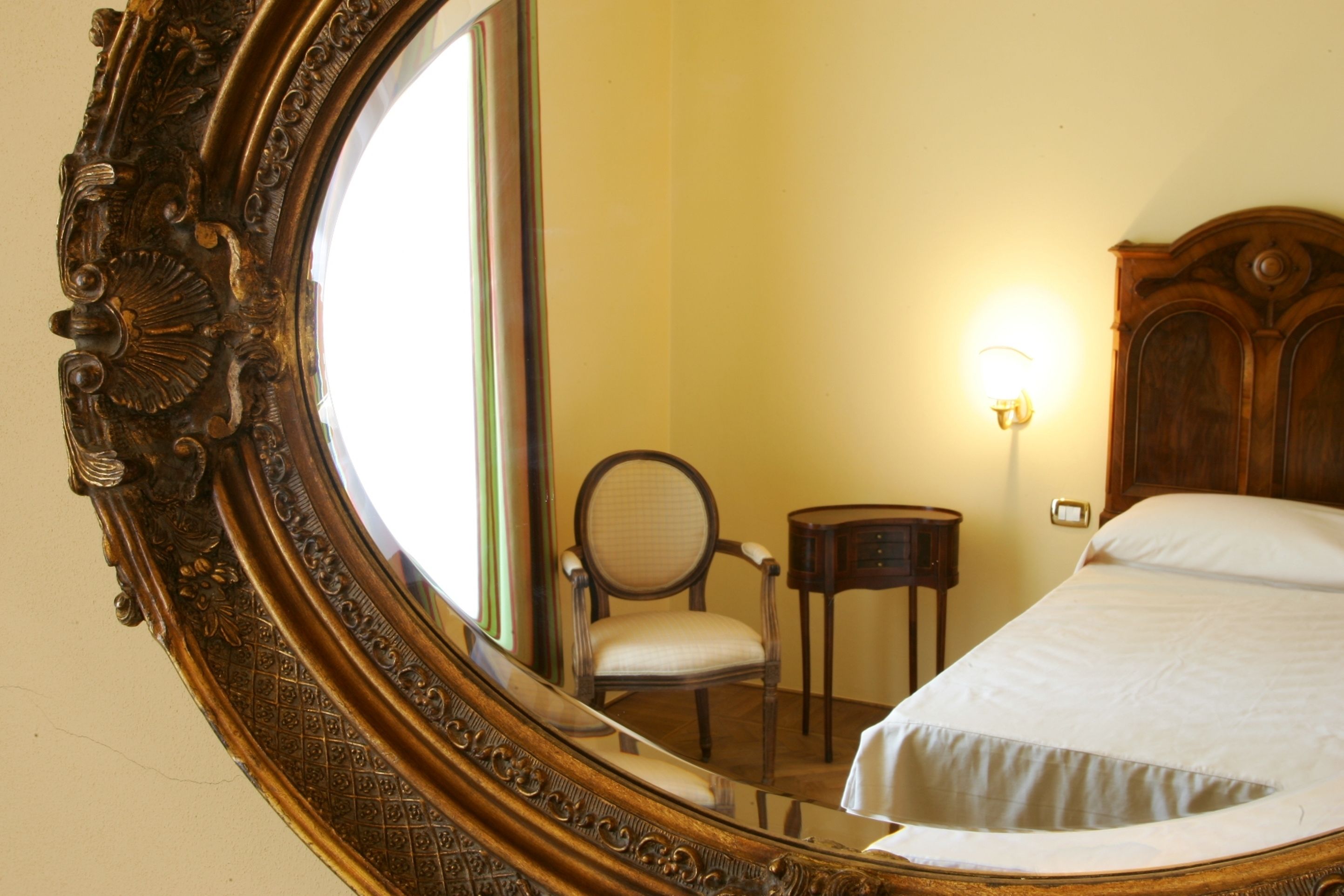 A bedroom with a large ornate mirror, a wooden headboard, a bedside table, and a chair.