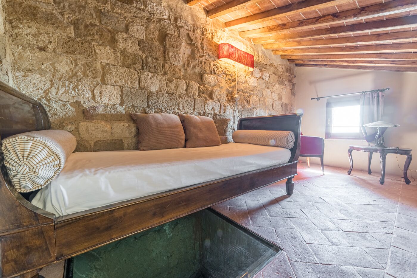 A rustic room with a stone wall, a wooden bed with a mattress and pillows, a small table, and a chair.