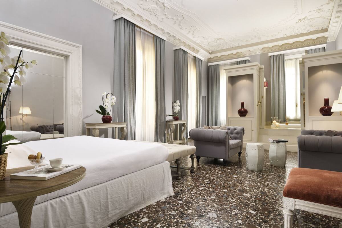 Habitación de hotel con una cama grande, dos sillones, una mesita con un florero y un espejo.