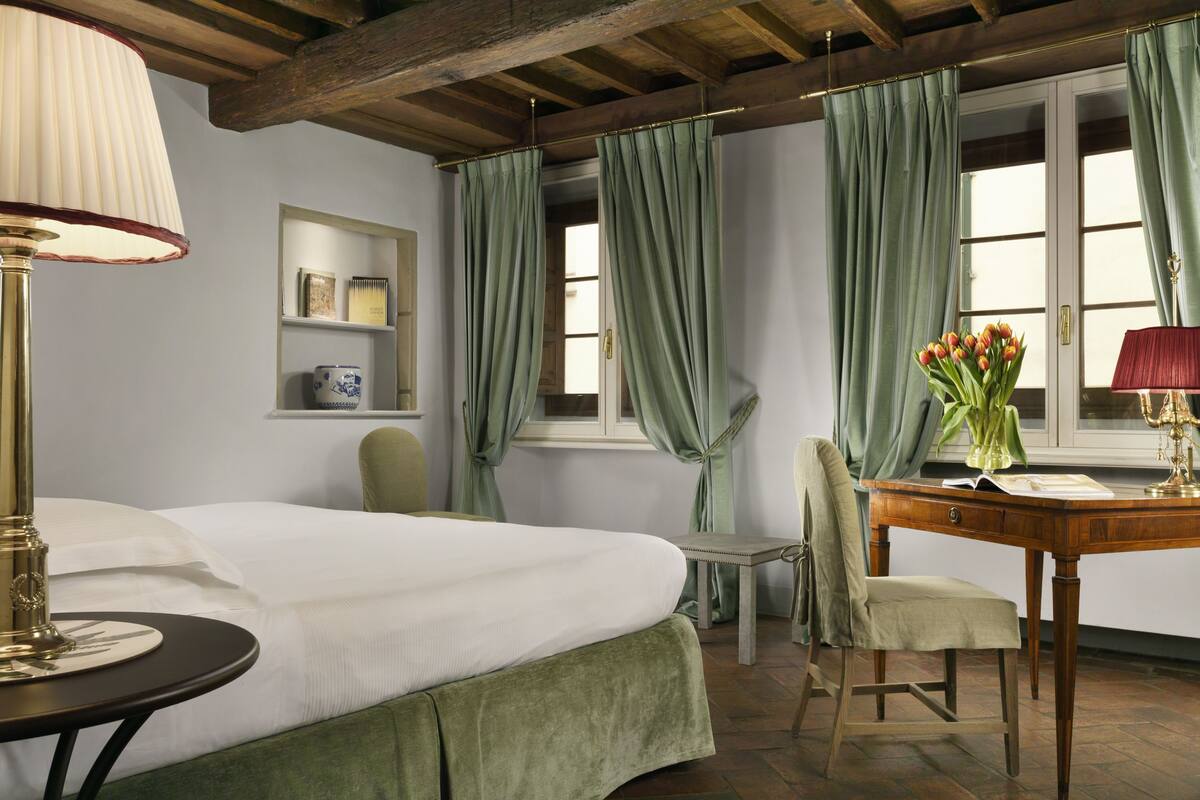 Una habitación con cama, escritorio, silla, lámpara, un jarrón con flores y una ventana con cortinas.