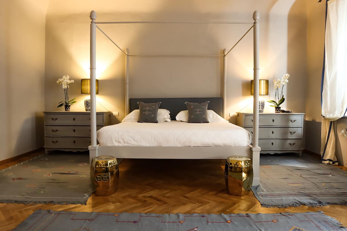 Una cama con dosel, ropa de cama blanca, dos mesitas de noche con lámparas y jarrones decorativos a cada lado.