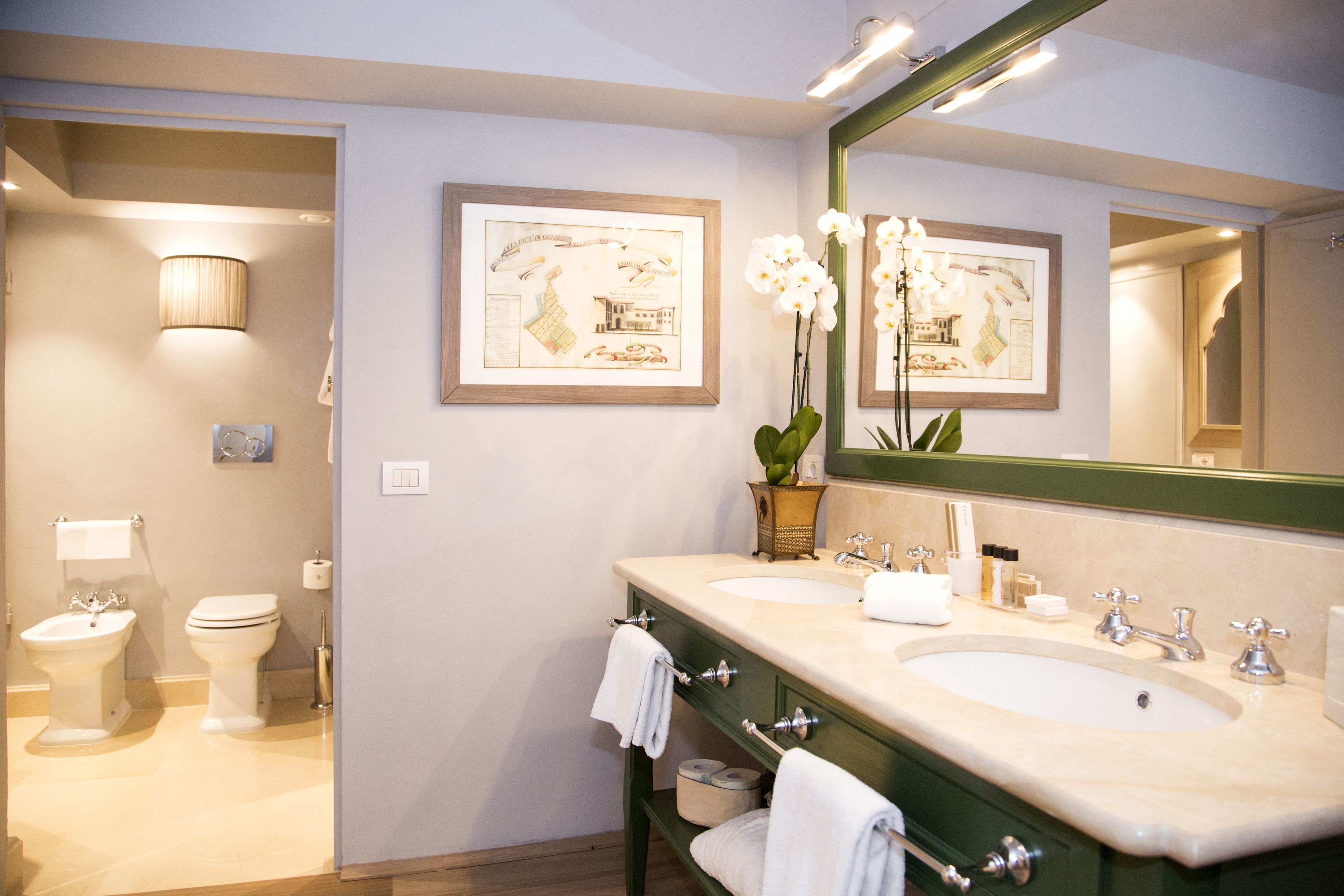 A bathroom with two sinks, a toilet, a mirror, and framed artwork on the wall.