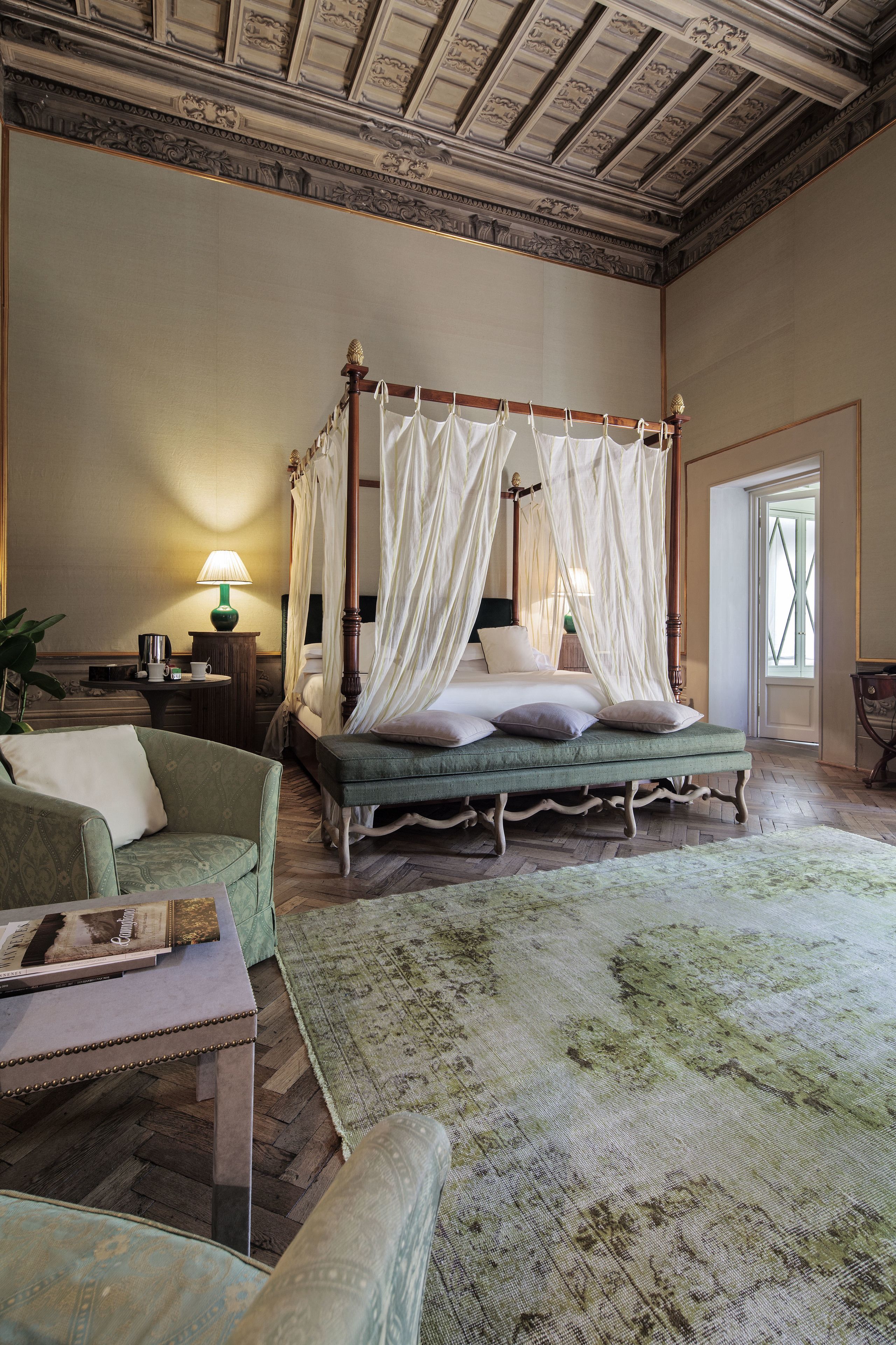 Un lit à baldaquin à quatre poteaux, doté d’une tête de lit recouverte d’un tissu vert, une table de chevet avec une lampe et une grande fenêtre offrant une belle vue.