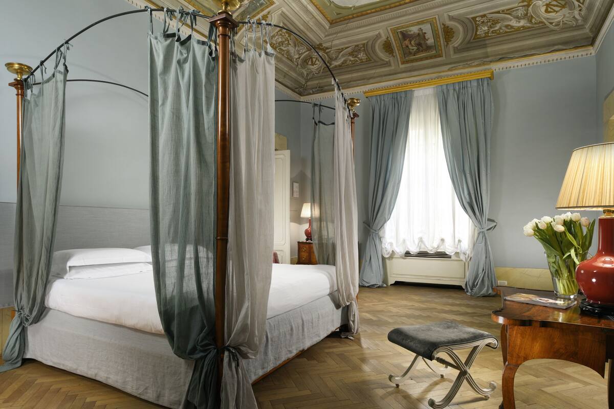 Una cama con dosel, cortinas traslúcidas, un banco de madera, una mesita de noche y un florero con flores.