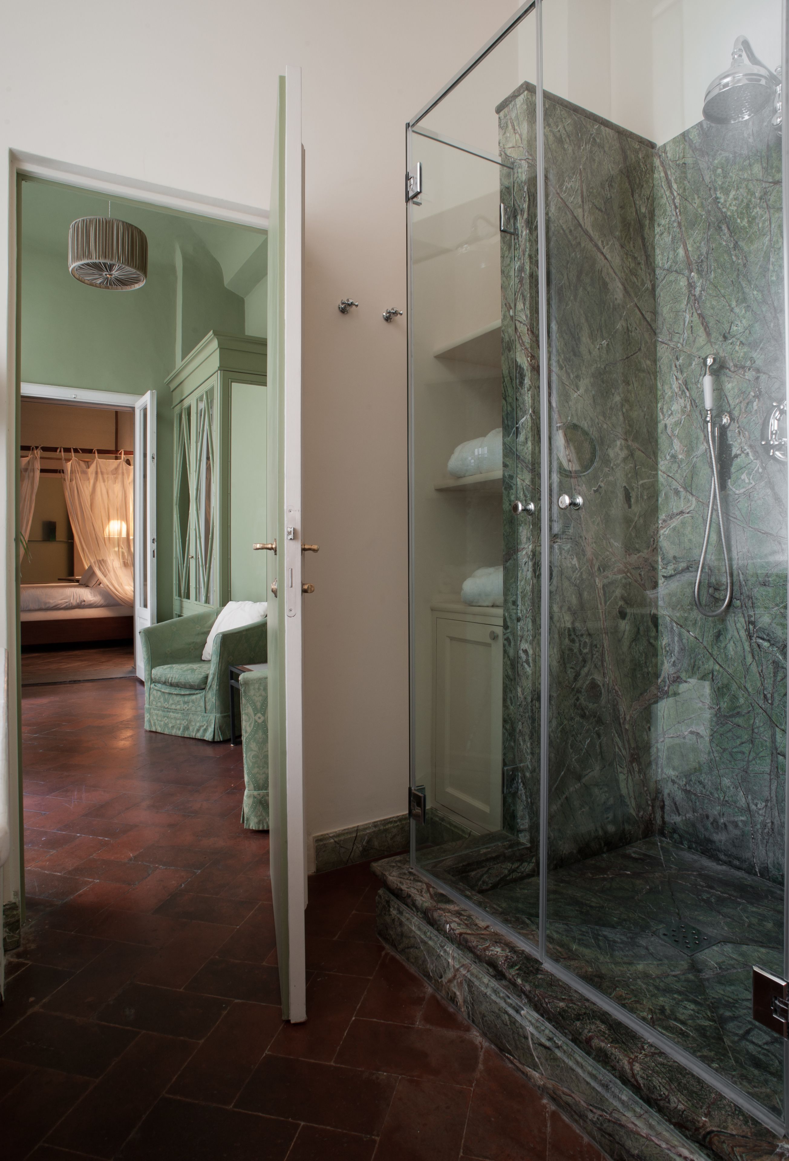 A modern bathroom with a glass-enclosed shower, marble walls, and a visible bed in the adjacent room.