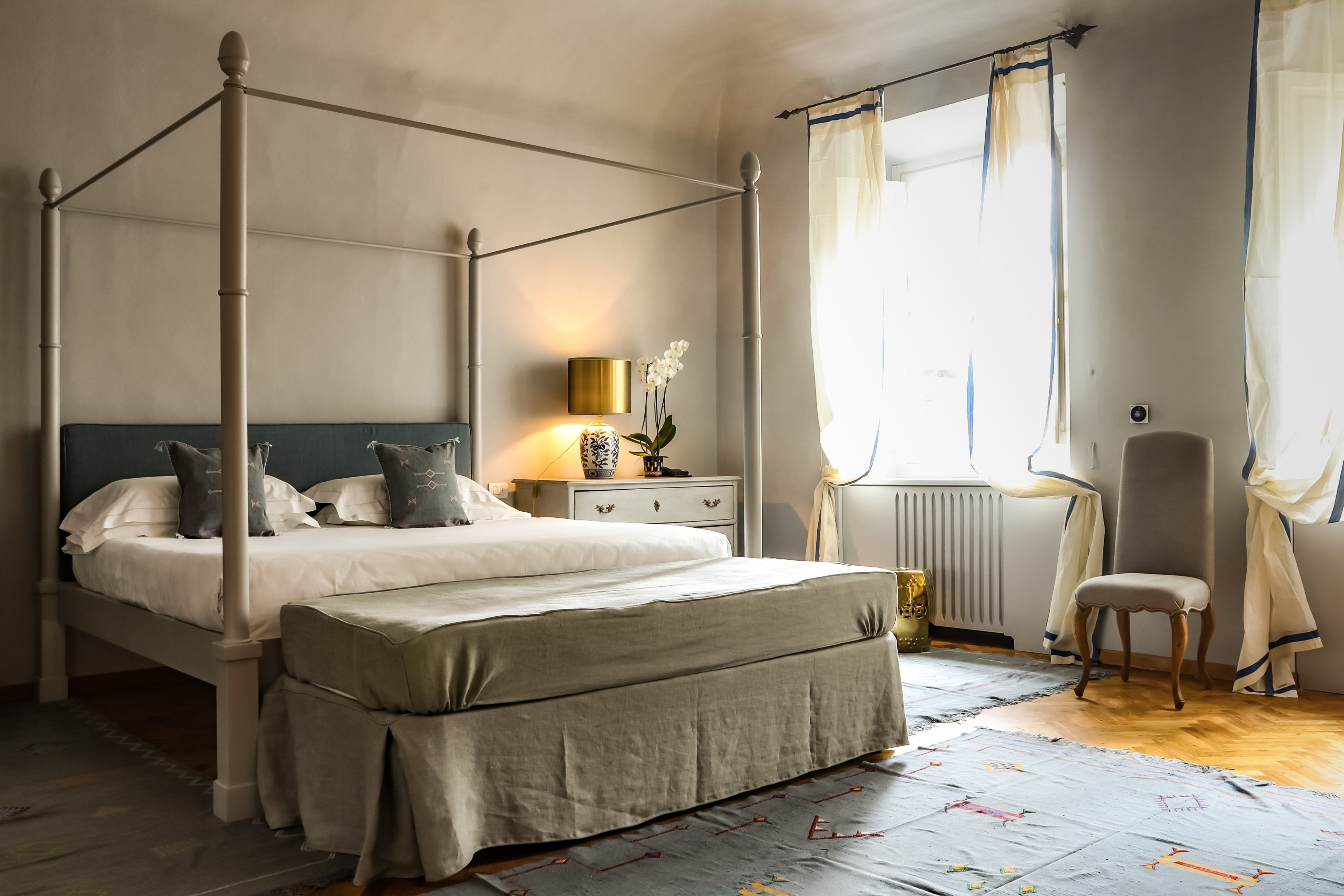 A four-poster bed with a matching bench, a bedside table with a lamp, and a chair near a window with sheer curtains.