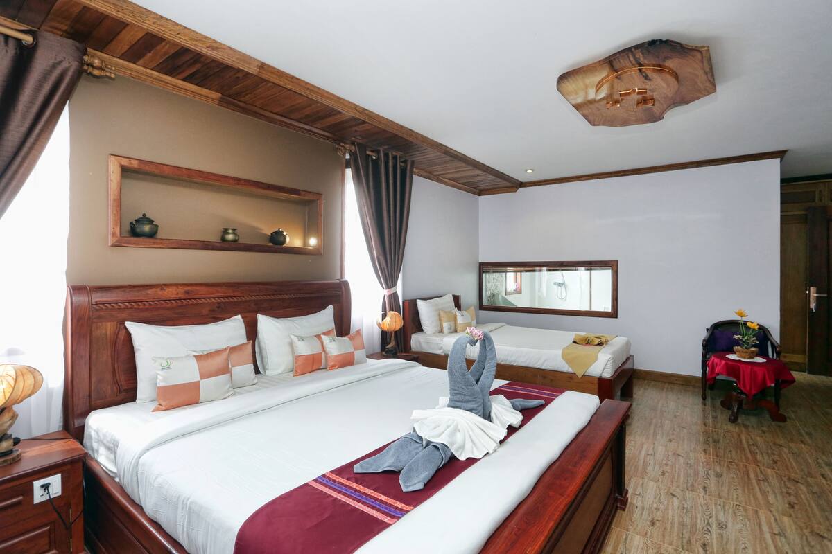 A hotel room with a large bed, a wooden headboard, a bedside table, a mirror, a window, and a red tablecloth on a chair.