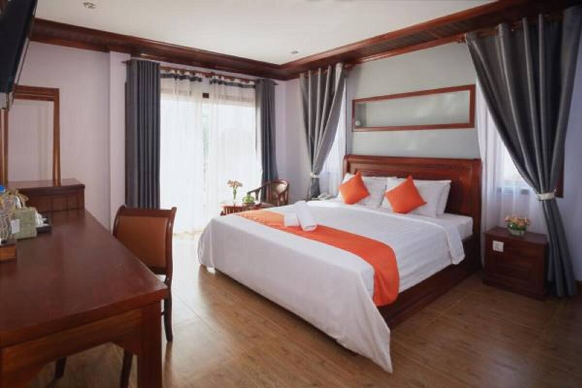 A hotel room with a wooden bed, white bedding, orange pillows, a wooden nightstand, a desk, and a chair.