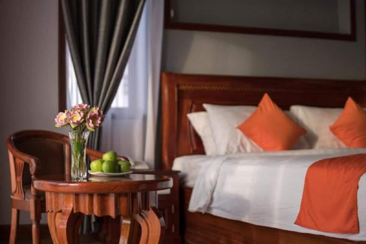 A hotel room with a wooden bed, white bedding, and orange pillows. There is a wooden chair and a table with a vase of flowers and a plate of fruit.
