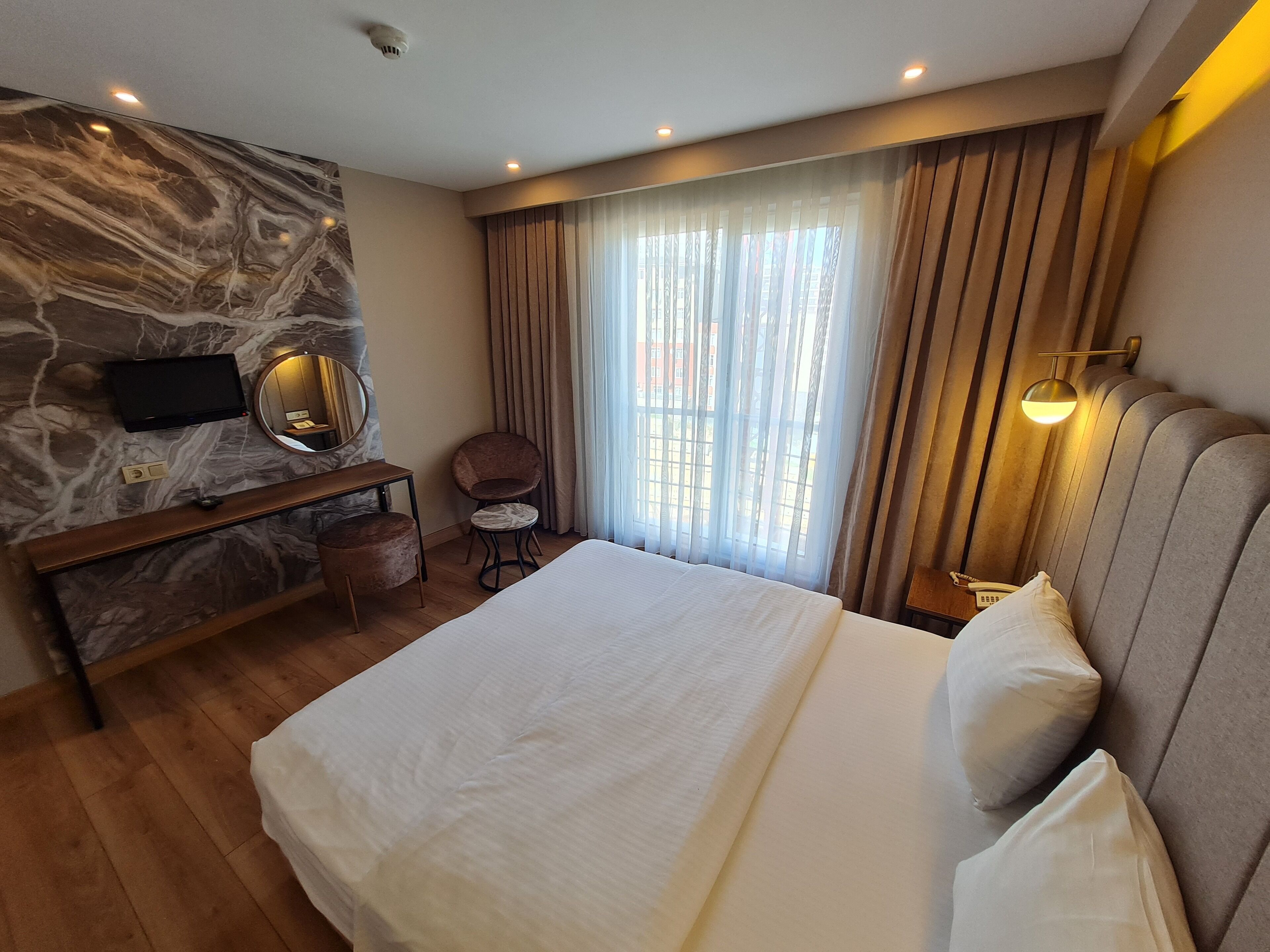 A hotel room with a marble wall, a bed with white linens, a round mirror, a small round table, a chair, and a window with curtains.