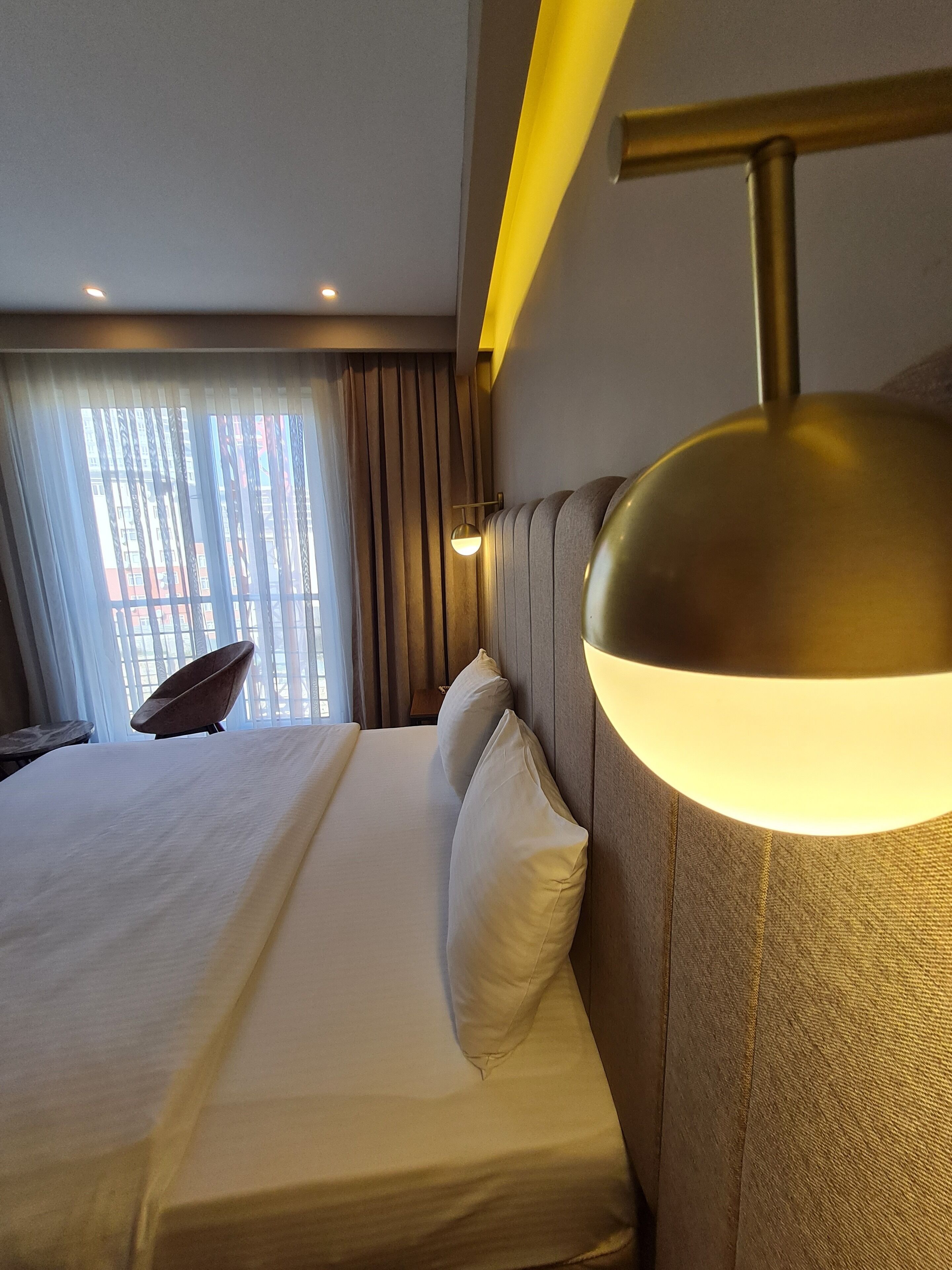 Une chambre d’hôtel avec un lit bien fait, une lampe fixée au mur et une vue sur l’extérieur par une fenêtre ornée de rideaux.